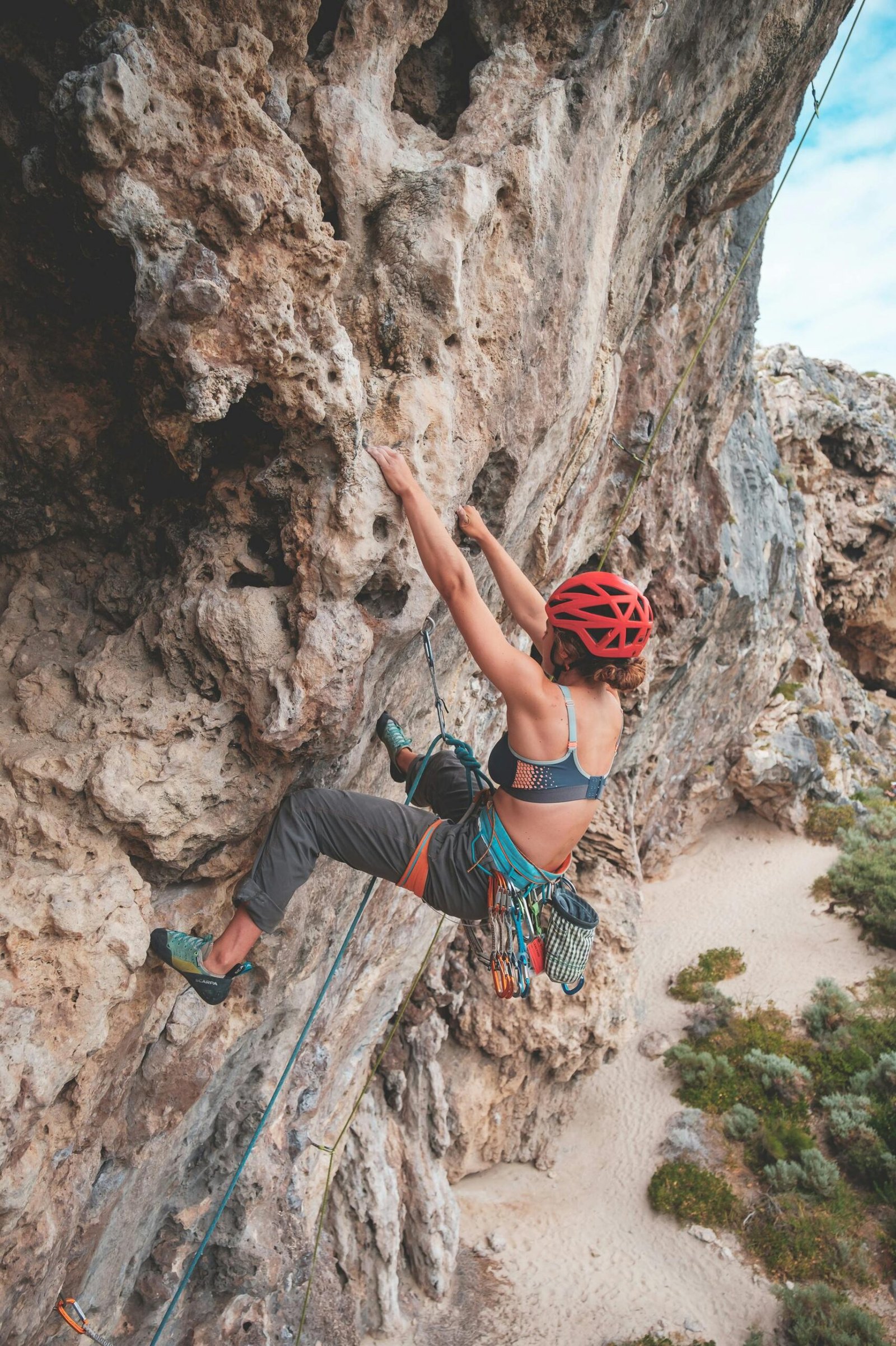 A woman rock climbing on a limestone cliff, wearing a helmet and climbing gear, secured by a rope. Sandy ground and bushes are visible below.